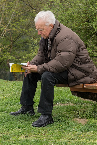 To learn the benefits of financial literary education, a retired man with short white hair and a serious look on his face, sits on a park bench wearing a brown puffy jacket and black loose pants reading an educational booklet on savings and retirement.