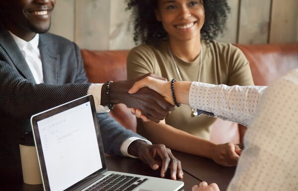 Happy young african american couple making deal handshaking caucasian insurance broker in cafe, black satisfied customer and realtor or sales person shaking hands at meeting in office with laptop