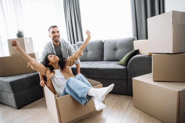 Happy multiracial couple is having fun with cardboard boxes in their new house at moving day. Afro american girl and caucasian guy among the boxes in his new house, guy pushes the girl while she sits