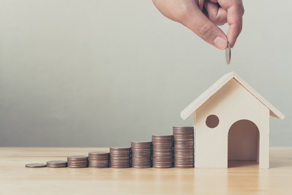 Hand putting a coin in a small wooden house; stacks of coins, from short to tall, are stacked beside the house