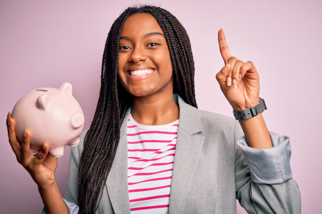 Young african american business woman saving money on piggy bank over isolated background surprised with an idea or question pointing finger with happy face, number one